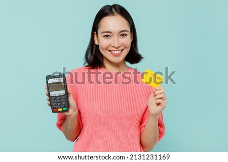 Young smiling fun woman of Asian ethnicity 20s in pink sweater hold wireless modern bank payment terminal to process acquire credit card payments isolated on pastel plain light blue background studio Royalty-Free Stock Photo #2131231169