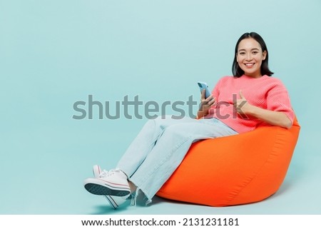 Full body young smiling happy woman of Asian ethnicity 20s wearing pink sweater sit in bag chair hold in hand use mobile cell phone show thumb up gesture isolated on pastel plain light blue background Royalty-Free Stock Photo #2131231181