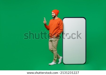 Full body young smiling man 20s wear orange sweatshirt hat stand near big blank screen mobile cell phone with workspace copy space mockup area use smartphone isolated on plain green background studio