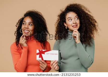 Two dreamful fun young curly black women friends 20s wear casual shirts clothes hold gift certificate coupon voucher card for store look aside isolated on plain pastel beige background studio portrait