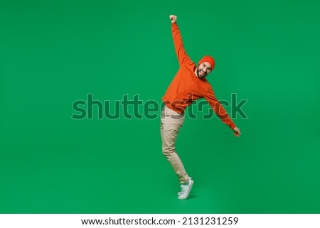 Full body young fun smiling handsome attractive happy cool man 20s wearing orange sweatshirt hat leaning back with putstretched hands stand on toes isolated on plain green background studio portrait.