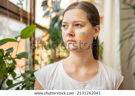 A sad young Ukrainian girl with the flag of Ukraine on her face is looking through window. The concept of participation of the Ukrainian people in the war with Russia. Not war concept. Quality photo