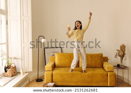 Beautiful energetic brown-haired woman is dancing on couch to music from her phone at home. Young model spends time alone on sunny day, wearing yellow blouse and white pants. Holiday life concept Royalty-Free Stock Photo #2131262681
