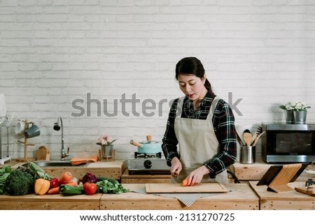 Picture of happy woman preparing diet healthy meal while slicing carrot by using sharp knife at home kitchen. beautiful lady in apron cutting fresh vegetables in wooden cooking place interior.