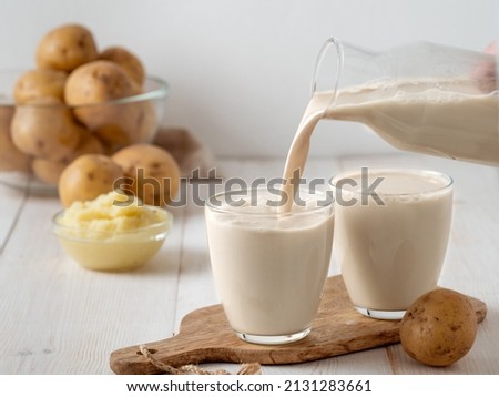 Potato milk pouring into glass on white wooden background. Pouring vegan milk in glass, with potato puree and potato tubers on background. Copy space. Home made potato milk made from boiled potatoes Royalty-Free Stock Photo #2131283661