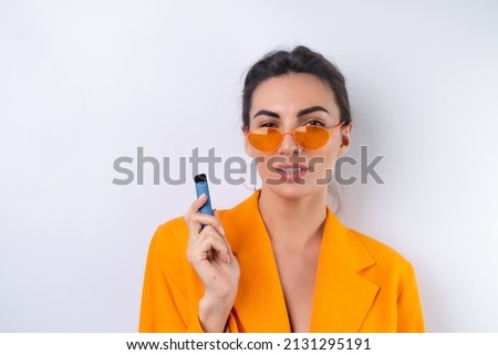 A young woman in trendy stylish glasses and a bright orange oversized jacket on a white background smokes an electronic cigarette