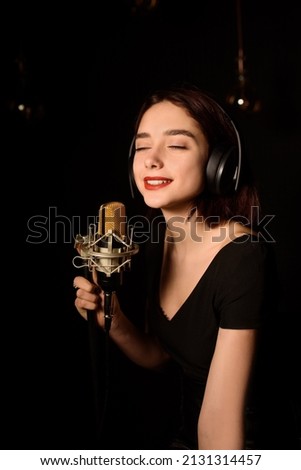 Portrait of young woman with headphones is recording a song in an audio recording studio on black background.

