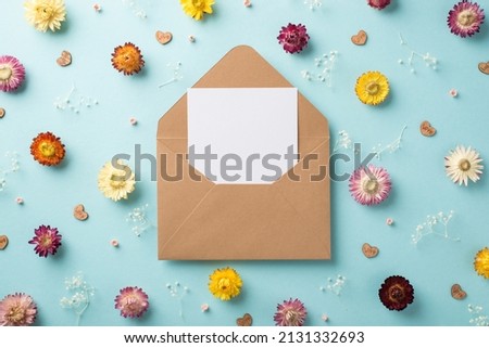 Top view photo of the big open craft envelope with paper card inside and many different cute flowers white gypsophila branches and wooden confetti in shape of hearts scattered on the pastel blue backg