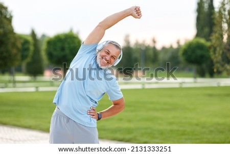 Staying active after retirement. Happy joyful mature retired sportsman wearing headphones and sportswear doing side stretching exercises with arm over his head, exercising outside in city park Royalty-Free Stock Photo #2131333251
