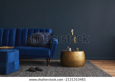 Stylish modern living room interior design with glamour blue velvet sofa, pouf, golden metal side table and modern home accessories. Dark blue wall. Template. Copy space.
