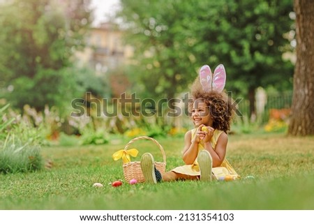 Little Black girl wear bunny ears and gathering Easter eggs on Easter egg hunt in garden Royalty-Free Stock Photo #2131354103