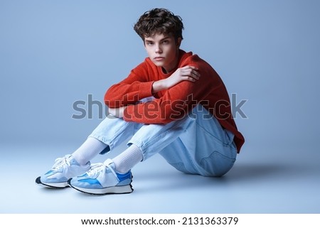 Handsome guy model in a red sweater, light jeans and sneakers poses sitting on the floor in the studio. Men's youth fashion.  Royalty-Free Stock Photo #2131363379