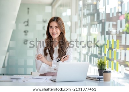 Beautiful smiling young Asian businesswoman sitting with arms crossed looking at camera in the office.