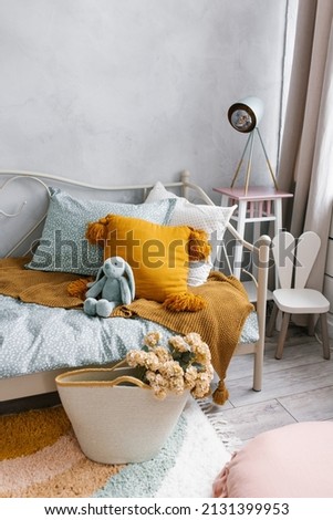 A bed in the children's bedroom with a bunny toy and pillows. Next to the bed is a wicker basket with flowers as a decorative element. Modern interior of the children's room Royalty-Free Stock Photo #2131399953