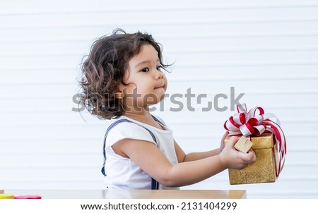 Portrait little caucasian curly hair girl wearing casual clothes, holding gift or present box with surprising face, while celebrating birthday or christmas at home. Kid and Education Concept