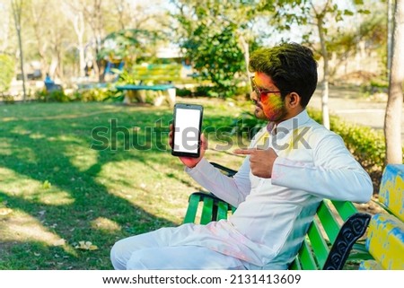 Festival and technology concept - indian young man playing holi colors and using phone or showing screen on camera