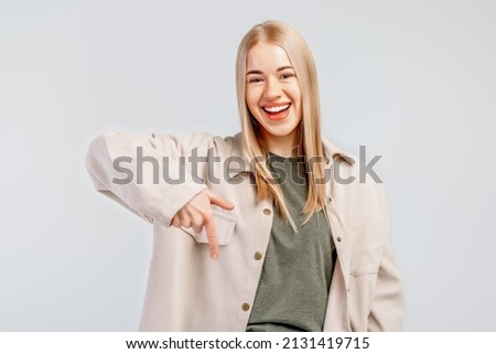 Portrait of smiling stylish blond girl in trendy shirt pointing finger down, showing promo deal, inviting to register, advertisement, standing over gray background