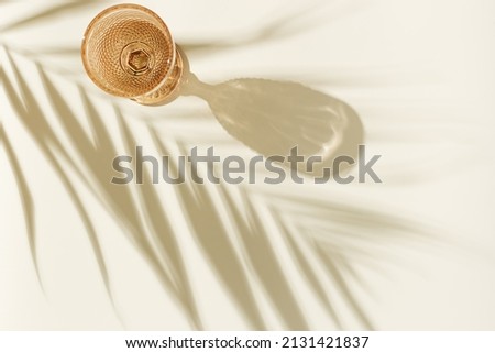 White wine glass peach color glass on beige background with palm leaf shadow, glare at sun. Minimal summer rest concept. Dry wine in colored glassware goblets style. Creative top view, pastel colors