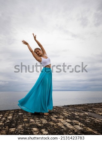 Beautiful woman practicing ballet pose. Young ballerina wearing long blue skirt and dancing. Outdoor ballet practice. Slim body. Cloudy sky background. Copy space. Bali, Indonesia