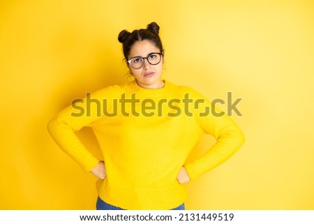 Young beautiful woman wearing casual sweater over isolated yellow background skeptic and nervous, disapproving expression on face with arms in waist