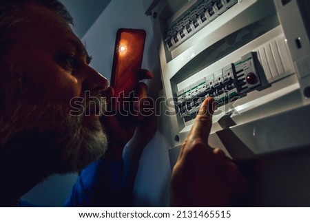 Energy crisis. Man in complete darkness holding a phone to investigate a home fuse box during a power outage. Blackout concept. Royalty-Free Stock Photo #2131465515