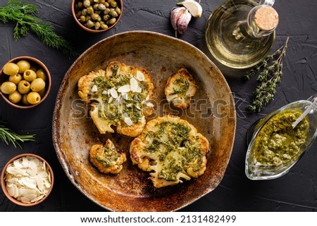 Cauliflower steak with spices lies in a frying pan. Olive oil, chimichurri sauce, capers, olives, herbs, various spices side by side. Dark background. Vegetarian food. Royalty-Free Stock Photo #2131482499