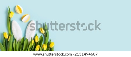 Yellow tulips, paper crafting eggs and white fluffy rabbit ears on blue background, easter and spring concept, top view, copy space