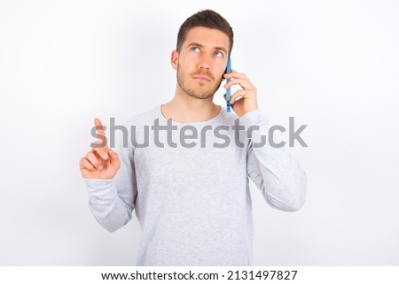 young caucasian man wearing grey sweater over white background speaks on mobile phone spends free time indoors calls to friend.