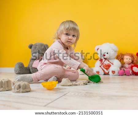 A two-year-old girl in a pink suit is sitting on the floor in the playroom and playing with beige kinetic sand