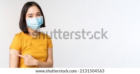 Covid-19, vaccination and healthcare concept. Portrait of cute asian girl in medical mask, has band aid on shoulder after coronavirus vaccine, standing over white background