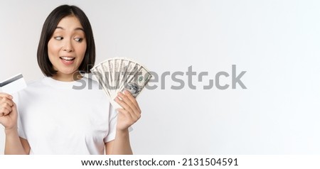 Portrait of asian woman holding money, dollars and credit card, looking impressed and amazed, standing in tshirt over white background