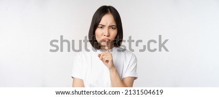 Angry asian girl shushing, keep quiet, taboo silence gesture, press finger to lips and frowning, scolding for being too loud, standing over white background