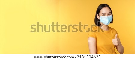 Vaccination from covid and health concept. Happy asian girl showing thumbs up, wearing medical mask, band aid on shoulder, got coronavirus vaccine shot, yellow background