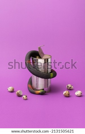 A rubber snake toy comes out of a metal can with egg shells around on a purple background. Minimal concept with copy spaceac. Side view.