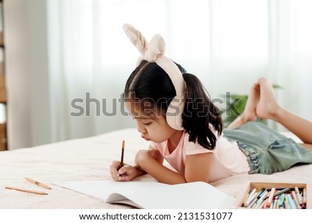4k 50fps Cute little girl having fun in cute kindergarten age. Use colored pencils to draw a picture on the paper. Happily put the rabbit headphones on the bed. Concept family and happy learning
