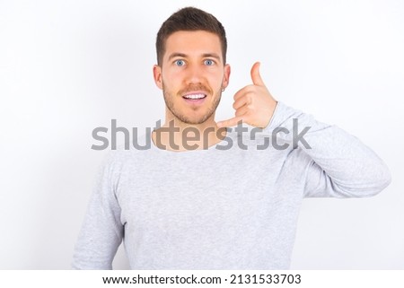 young caucasian man wearing grey sweater over white background makes phone gesture, says call me back again, has glad expression.