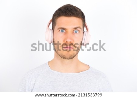 Serious displeased young caucasian man wearing grey sweater over white background looks puzzled at camera being angry wears stereo headphones listens music while walking at street