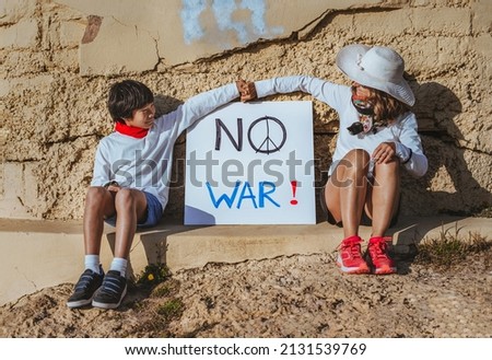 a mother and her asian son are sitting in front of a bunker and in the middle of them a sign that says "no war". they are holding hands. concept stop war and peace.