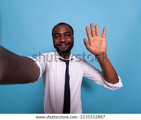 Smiling vlogger waving hello at camera in videocall conference on blue background. Wide angle pov of influencer taking a smartphone selfie doing hand gesture for social media.