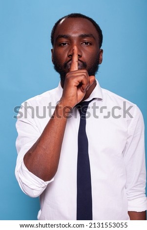 Portrait of african american man doing hush sign with index finger over lips on blue background. Calm businessman asking for silence doing shush hand gesture.