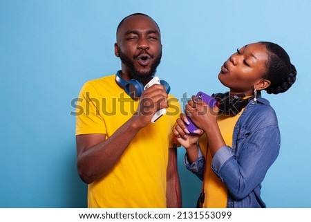 Trendy people doing karaoke with mobile phone as microphone in studio. Man and woman singing and having fun together with playlist music on headphones, enjoying mp3 sound on headset. Royalty-Free Stock Photo #2131553099