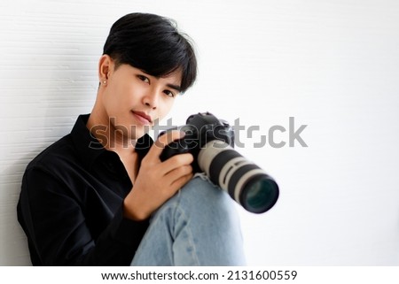 Asian handsome male model wearing casual black shirt with jeans, holding camera, sitting on white background in studio and posing while put his hand on hair