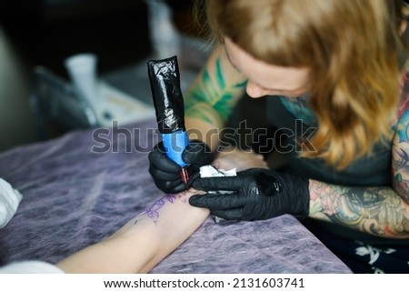 Cute European red-haired girl with tattoos makes a tattoo on woman arm. Woman creates a tattoo on a female hand by a professional tattoo artist