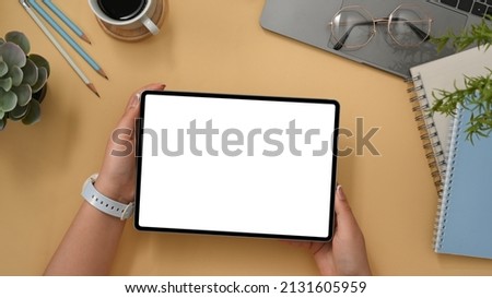 Comfortable workspace with office accessories, laptop and female holding tablet empty white screen mockup on beige background. clipping path, top view