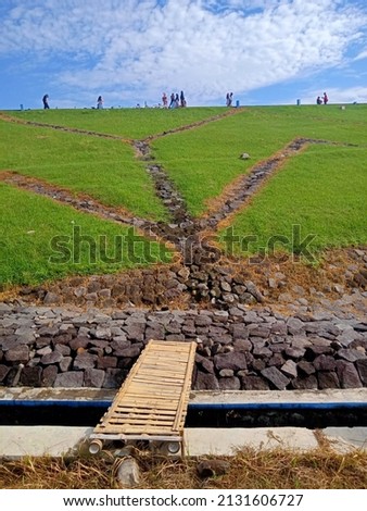 
Photo of a grassy hill with a bamboo bridge in the foreground and a bright blue sky in the background.