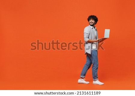 Full size body length side view young bearded Indian man 20s wears blue shirt hold use work on laptop pc computer typing browsing chatting send sms isolated on plain orange background studio portrait Royalty-Free Stock Photo #2131611189