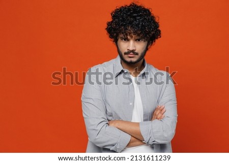 Sullen displeased dissatisfied gloomy young bearded Indian man 20s years old wears blue shirt hold hands crossed isolated on plain orange background studio portrait. People emotions lifestyle concept Royalty-Free Stock Photo #2131611239