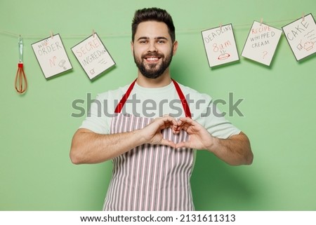 Young happy male chef confectioner baker man 20s in striped apron showing shape heart with hands heart-shape sign isolated on plain pastel light green background studio portrait. Cooking food concept