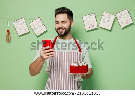 Young happy male chef confectioner baker man in striped apron hold birthday sweet dessert cake mobile cell phone isolated on plain pastel light green background studio portrait. Cooking food concept.