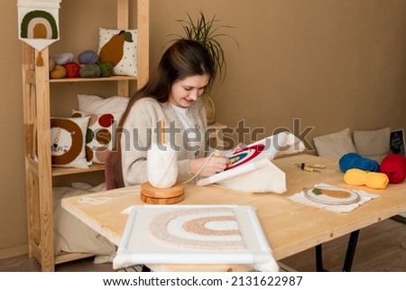 Punch needle embroidery. Young woman pushing the punchneedle straight down into the foundation fabric in workshop. Female embroidering handmade picture rainbow on canvas. Handicraft, hobby, diy Royalty-Free Stock Photo #2131622987